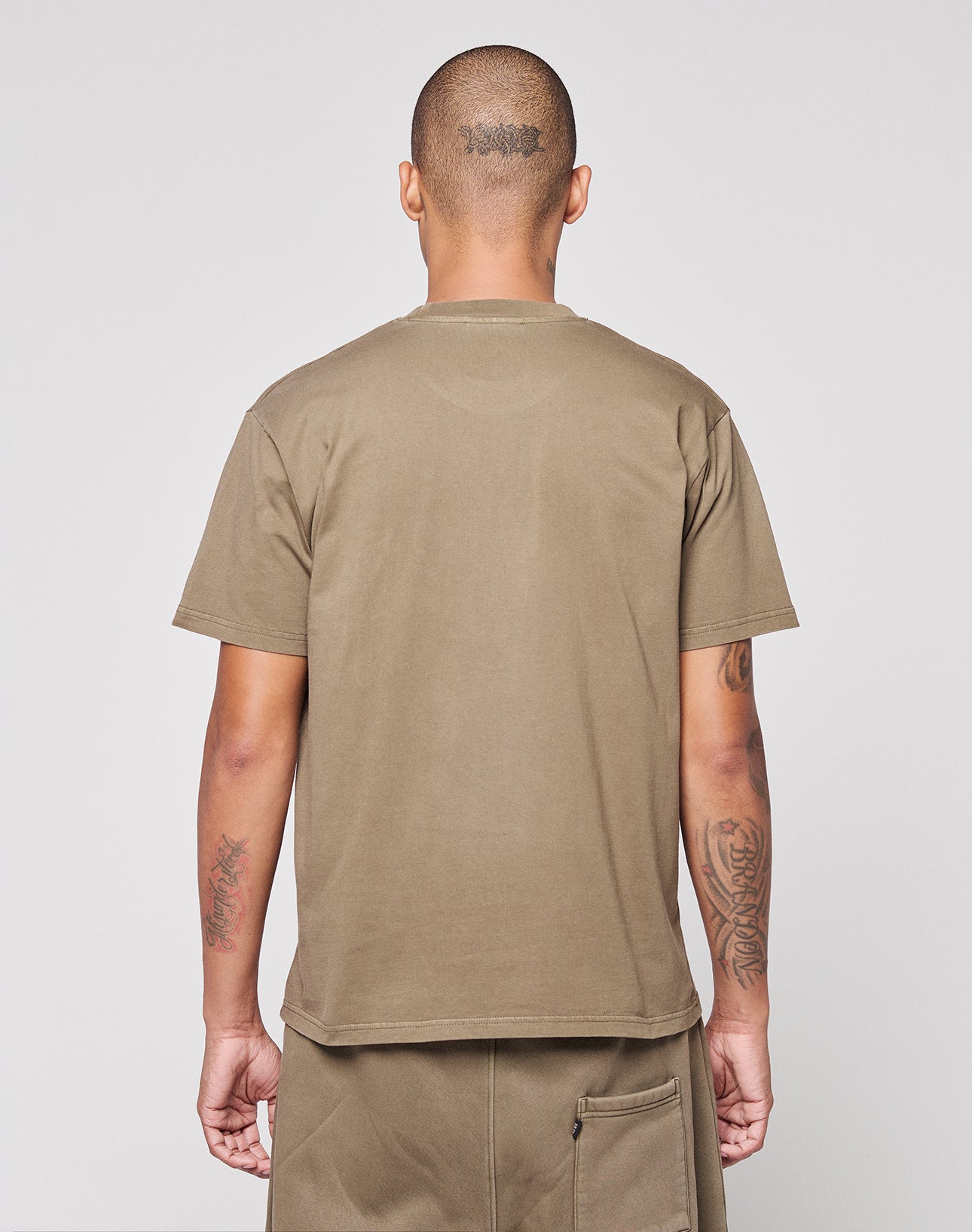 A person with a shaved head and back tattoos wears the LIVE FAST DIE YOUNG Live Fast Rhinestone Tee in olive-green oversize fit, paired with matching shorts, standing with their back to the camera against a plain light background.