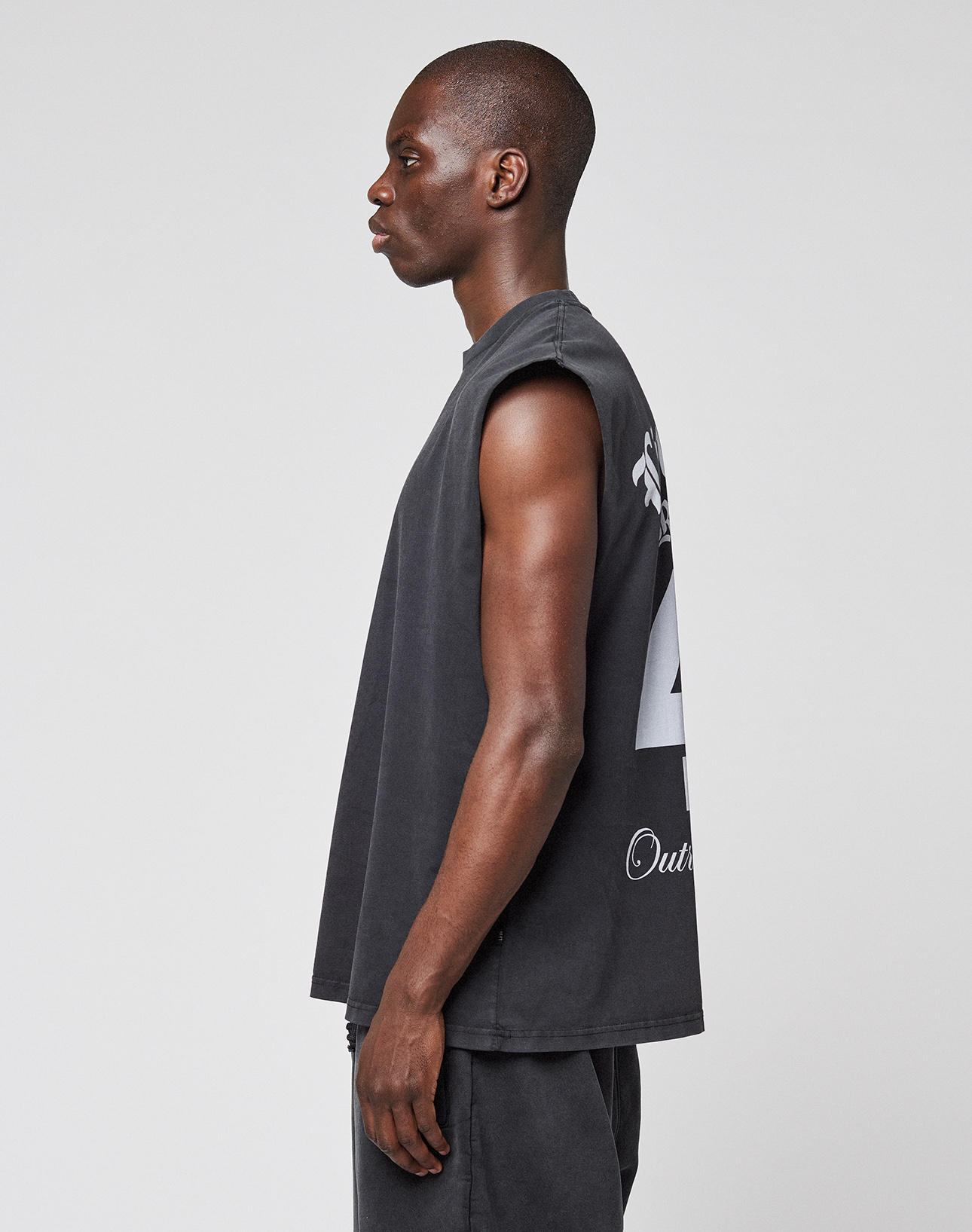 A person stands in profile against a plain background, wearing an oversize fit LIVE FAST DIE YOUNG LFDY Run Club Muscle Shirt—sleeveless and dark gray with large white back graphics—and matching dark pants.