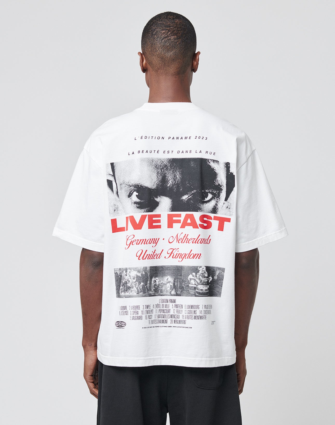 A person wears the LFDY Cinema Tee by LIVE FAST DIE YOUNG—an oversized white cotton T-shirt with a black-and-white face print, bold red LIVE FAST text, and detailed back design, photographed from behind on a plain background.