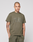 A man with neck and arm tattoos wears an oversized olive green LIVE FAST DIE YOUNG Live Fast Rhinestone Tee and matching pants, standing against a plain light gray background with a neutral expression and hands in his pockets.