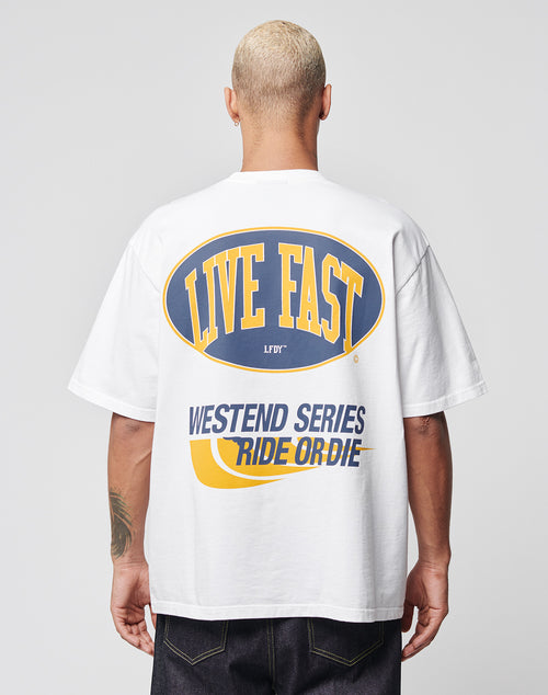 A person with short blond hair models the LFDY Westend Tee by LIVE FAST DIE YOUNG, an oversize white T-shirt with “LIVE FAST” in yellow and blue, and “WESTEND SERIES RIDE OR DIE” in blue and gold on the back. 100% Baumwolle.