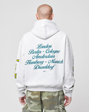 Wearing the LIVE FAST DIE YOUNG Rhine Area Hooded in oversize light gray cotton with a hood and seven European cities in blue script on the back, the person pairs it with camo pants, facing away against a plain background.