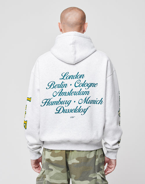 Wearing the LIVE FAST DIE YOUNG Rhine Area Hooded in oversize light gray cotton with a hood and seven European cities in blue script on the back, the person pairs it with camo pants, facing away against a plain background.