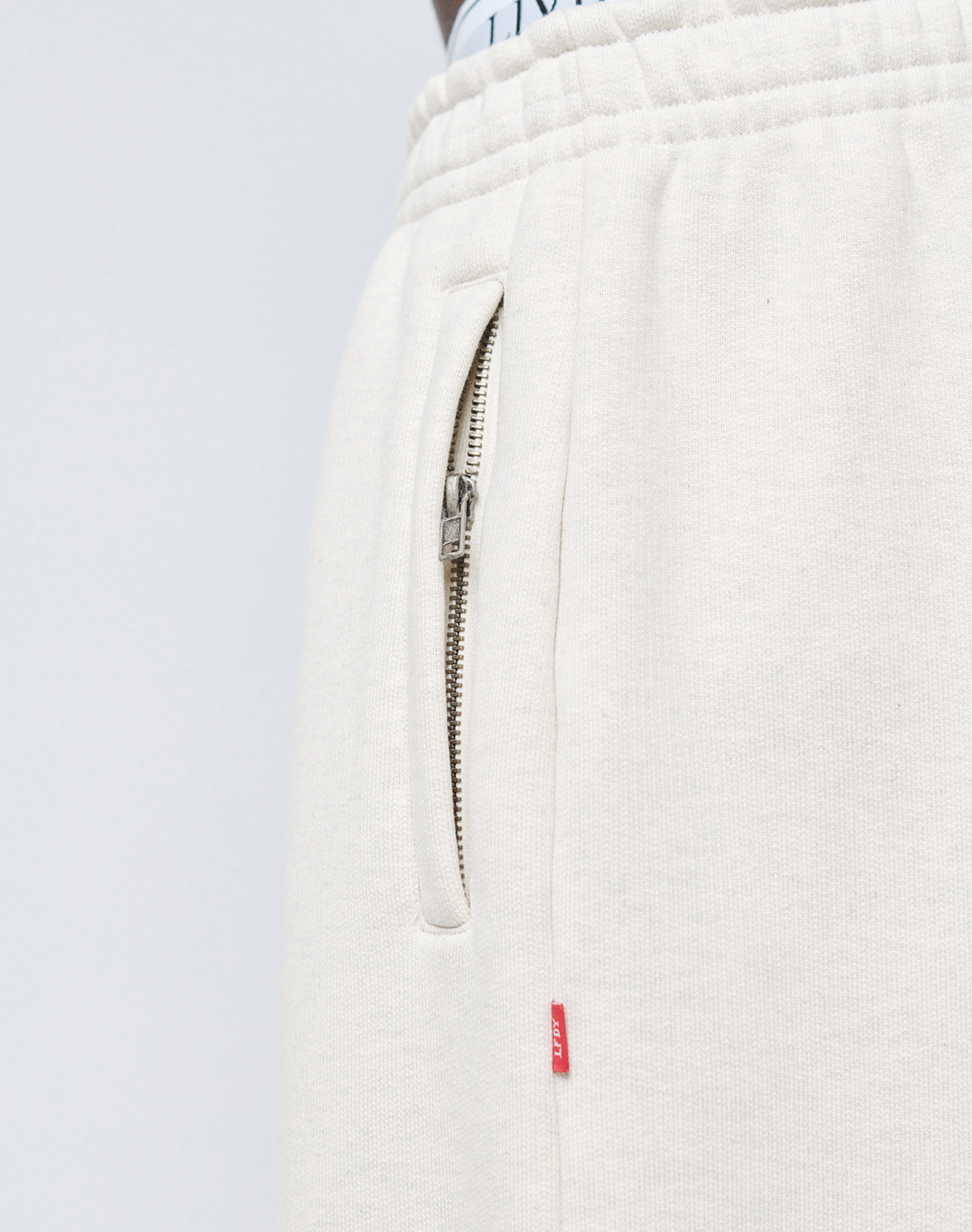 Close-up of cream-colored LFDY Heavyweight Loungepants by LIVE FAST DIE YOUNG, made from 650gsm cotton with an elastic waistband, metal zippered pocket, red fabric tag at the seam, and a white waistband with black text visible at the top.