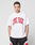 A person with short braided hair wears the LIVE FAST DIE YOUNG Live Fast College Tee, an oversize white t-shirt with bold red front letters, paired with black, white, and red shorts against a plain light gray background.