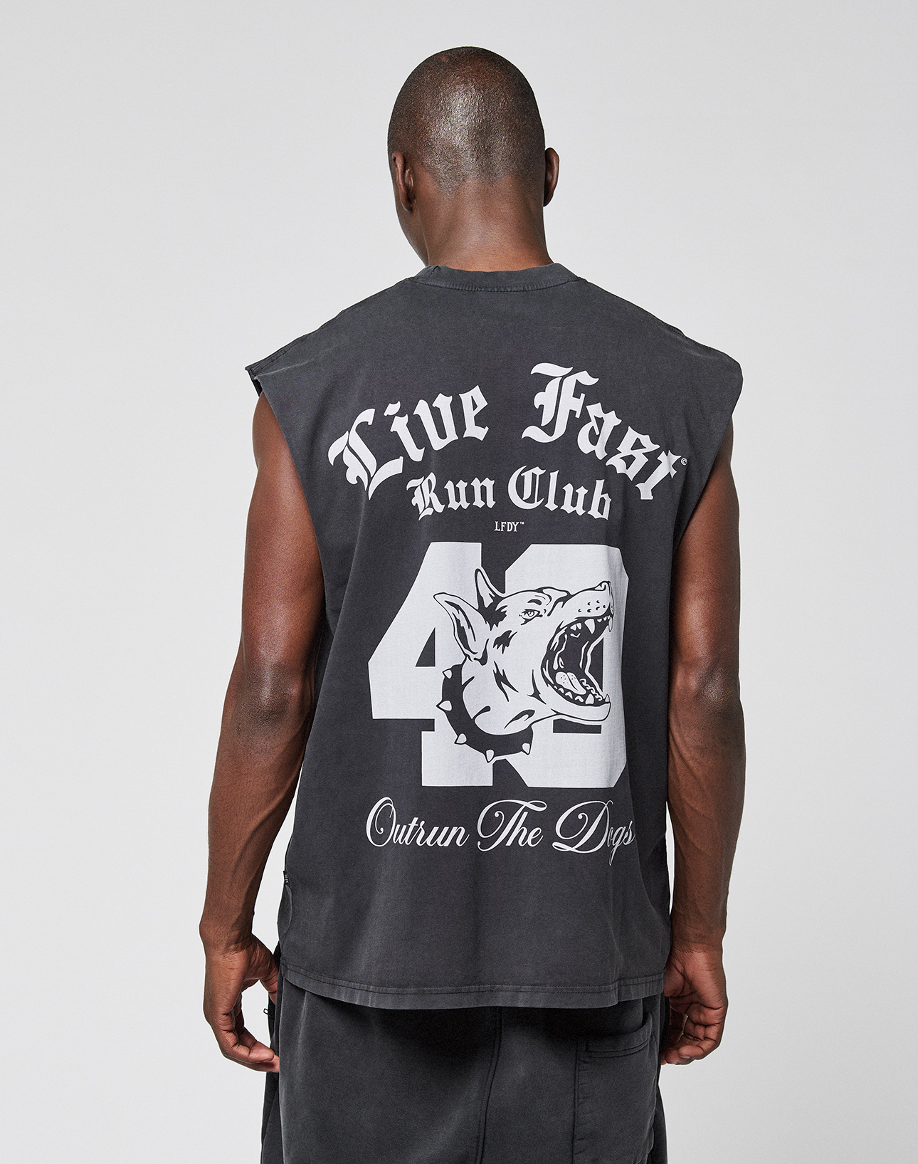 A man wears the LIVE FAST DIE YOUNG LFDY Run Club Muscle Shirt, a black vintage-look sleeveless top with an oversized fit, large 43, snarling dog graphic, "Live Fast Run Club" on the back, and "Outrun The Dogs" below.