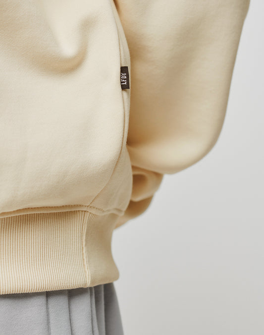 Close-up of the cream-colored Basic Blank Sweater by LIVE FAST DIE YOUNG, showing ribbed hem, detailed topstitching, and a small brown tag with white text. Worn over light grey pants against a plain light background.