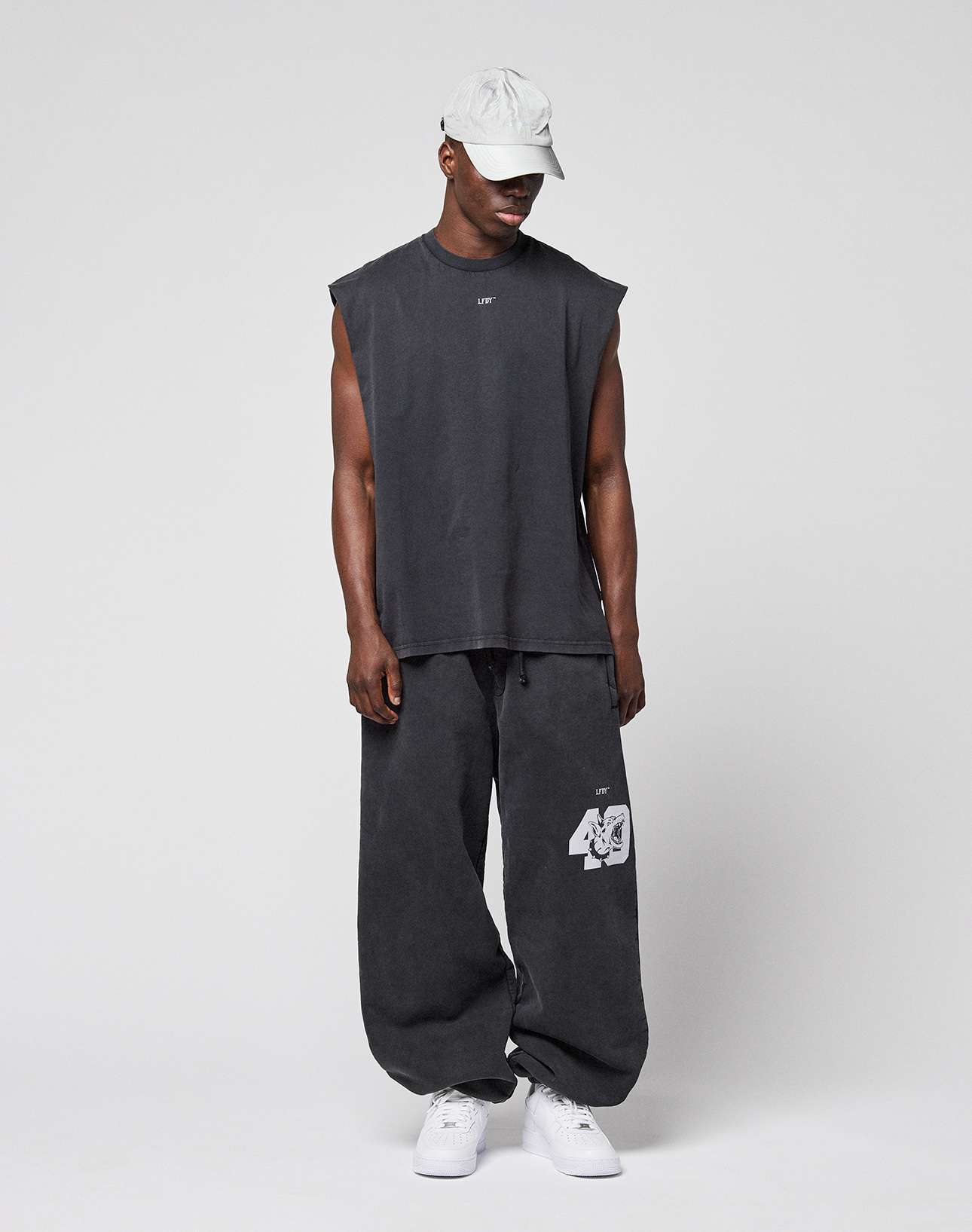 A man wears the LFDY Run Club Muscle Shirt by LIVE FAST DIE YOUNG, a sleeveless, oversized black top in 100% Baumwolle, with matching "49" print sweatpants and white sneakers, standing against a plain light gray background.
