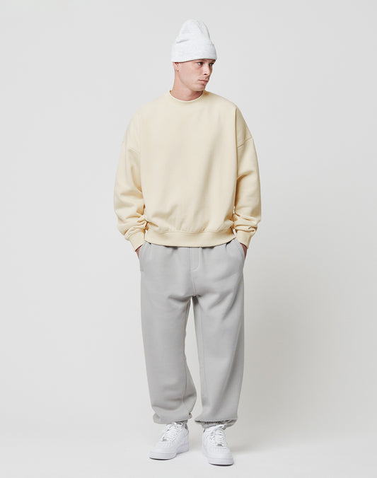 A person stands against a white background, wearing a LIVE FAST DIE YOUNG Basic Blank Sweater in cream, light gray sweatpants, white sneakers, and a white knit beanie—hands in pockets and gazing slightly to the side.