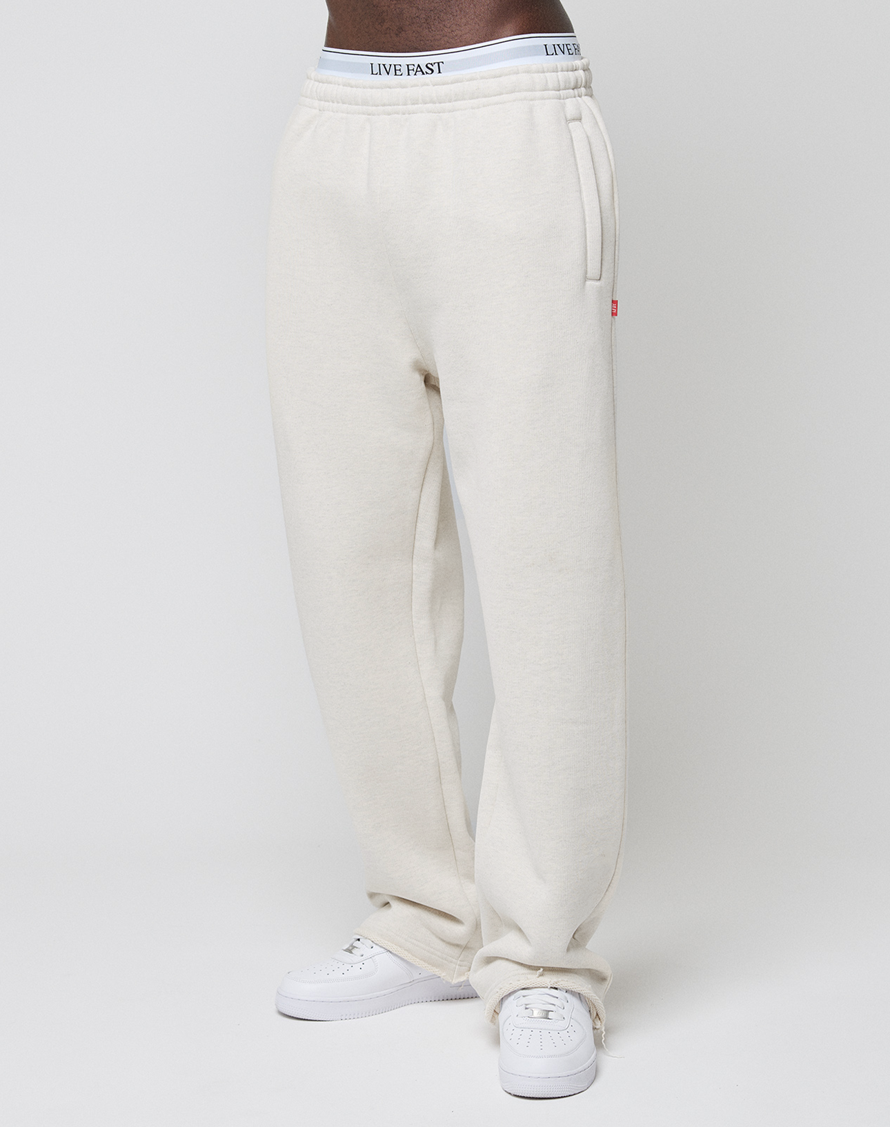 Person wearing off-white LFDY Heavyweight Loungepants by LIVE FAST DIE YOUNG with side pockets, relaxed wide fit, 650gsm heavyweight cotton, and a LIVE FAST waistband, paired with white sneakers against a plain light background.