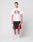 A man with braided hair stands against a plain background, wearing an oversize white LIVE FAST DIE YOUNG Live Fast College Tee with front print, paired with black and red athletic shorts, white socks, and white sneakers.
