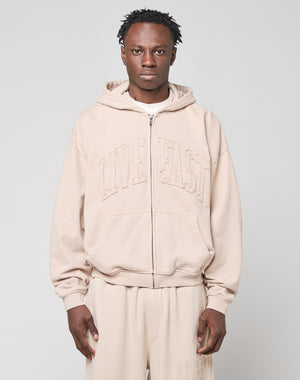 A person stands facing the camera against a plain light gray background, wearing the LIVE FAST DIE YOUNG Ripped College Hooded Zip in distressed beige Baumwolle. They have short, twisted hair and a neutral expression.