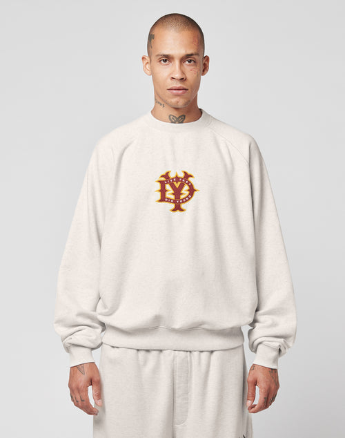 A person with a shaved head wears the LIVE FAST DIE YOUNG DY Sweater—an oversize, light gray sweatshirt with raglan sleeves and a red and gold interlocking letter emblem on the chest, paired with matching pants. The background is plain light gray.