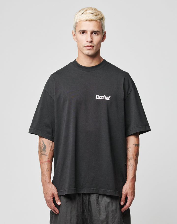 A young man with tattooed arms and short, bleached blond hair faces forward in a black LFDY Bulge Tee by LIVE FAST DIE YOUNG, featuring an oversize fit and live fast in white on the left chest. He has a neutral expression.