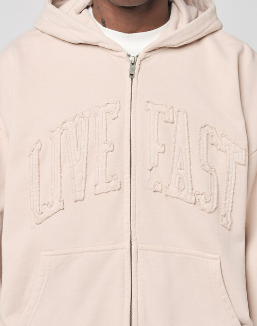 A person wears the LIVE FAST DIE YOUNG Ripped College Hooded Zip hoodie in light beige Baumwolle, featuring bold LIVE FAST embroidery. Only their chest and part of their face are visible.