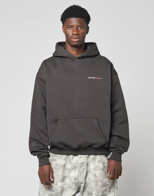 A man stands against a plain light background wearing an oversize fit black LIVE FAST DIE YOUNG LFDY F*ck Phones Hooded sweatshirt with Live Fast Archive text on the left chest and light gray camouflage pants.