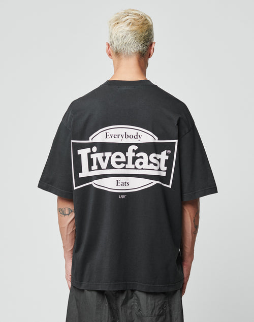 A person with short, bleached blonde hair is seen from behind wearing the LFDY Bulge Tee by LIVE FAST DIE YOUNG—an oversized black cotton T-shirt with bold front and back prints reading Everybody Eats Livefast in white and black.