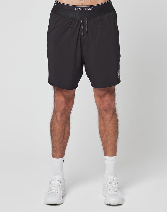 A person stands against a plain white background wearing LIVE FAST DIE YOUNG’s Live Fast Performance Shorts—black with LIVE FAST on the elastic waistband, regular fit—paired with white socks and sneakers. The photo is cropped at the waist and ankles.