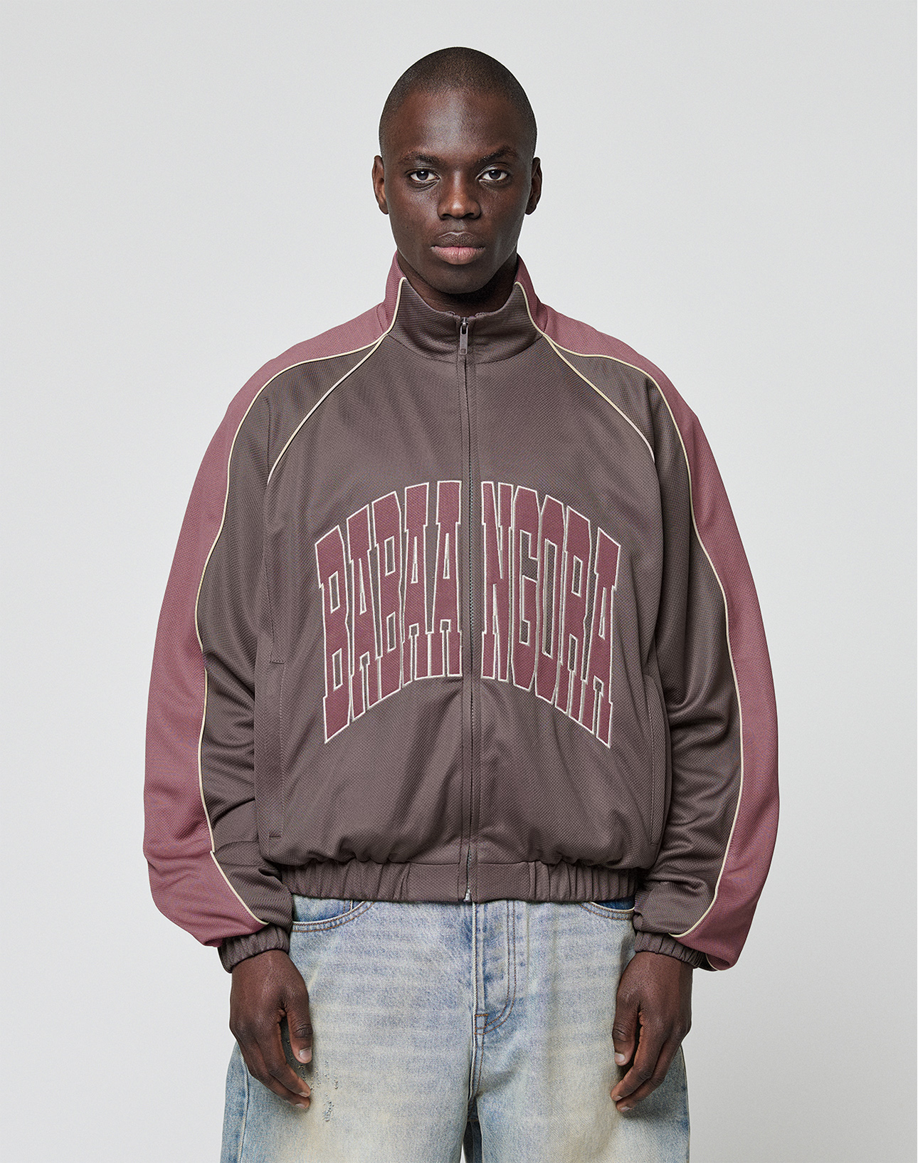 A man with short hair and light blue jeans faces forward, wearing the LIVE FAST DIE YOUNG LFDY x Baba Angora Trackjacket in brown and mauve, featuring bold BABA NIGORA lettering across the chest against a plain background.