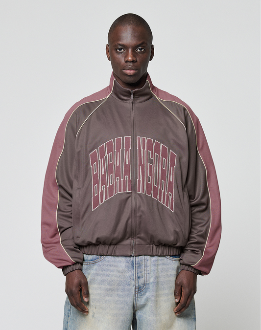 A man with short hair and light blue jeans faces forward, wearing the LIVE FAST DIE YOUNG LFDY x Baba Angora Trackjacket in brown and mauve, featuring bold BABA NIGORA lettering across the chest against a plain background.