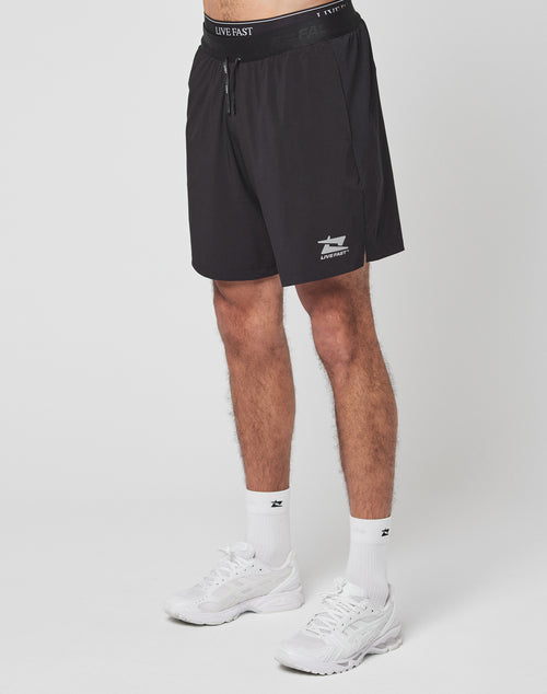 Someone wearing LIVE FAST DIE YOUNG Live Fast Performance Shorts in blackāwith a shark logo, reflektierender Print, and Elastikbundāplus white socks and sneakers stands against a plain white background. The upper body is not visible.