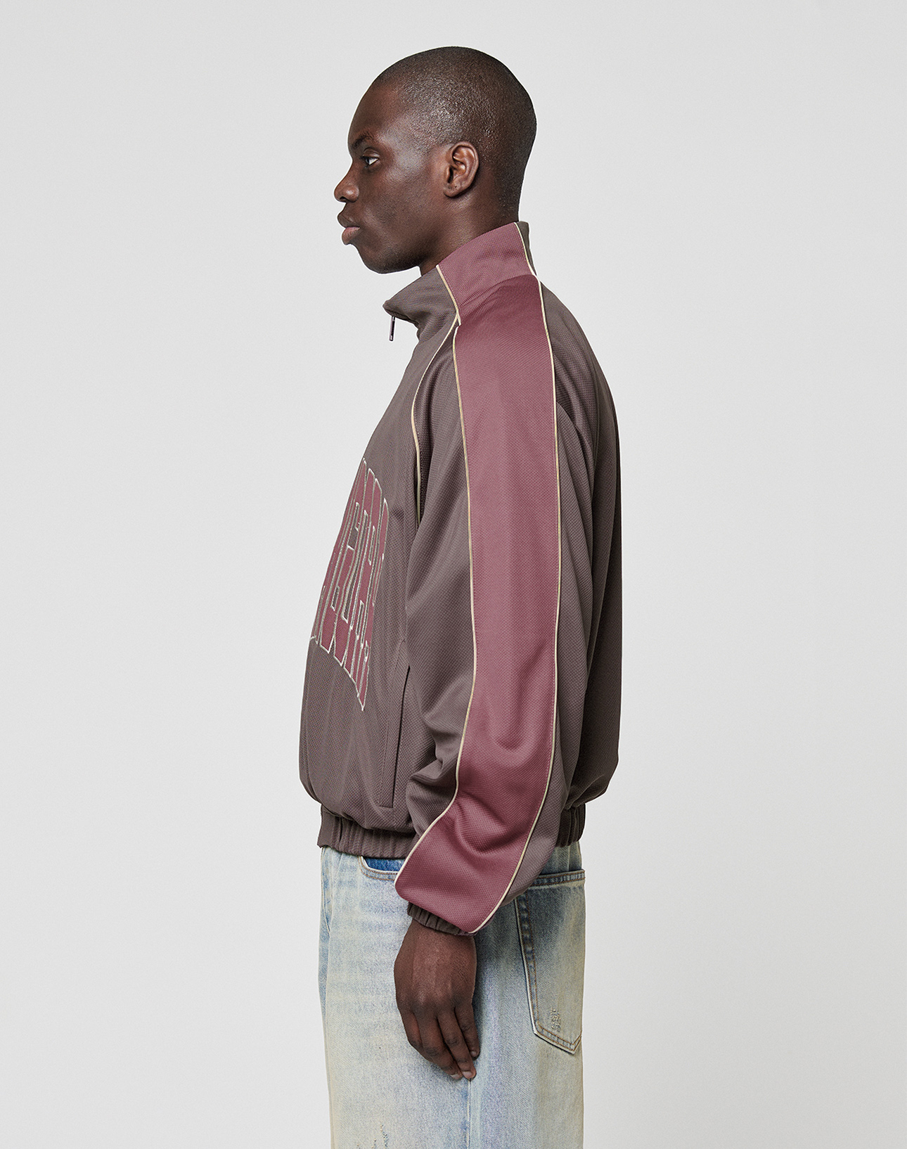 A person stands in profile against a plain background, wearing the LIVE FAST DIE YOUNG LFDY x Baba Angora Trackjacket—an oversize brown and maroon zip-up with Stick Applikation lettering—and light blue jeans.