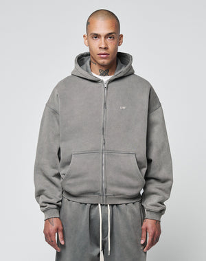 A person with a shaved head and neck tattoo wears the LIVE FAST DIE YOUNG Basic Hooded Zip and matching sweatpants, which have a logo print and lightly cropped fit, standing against a plain background facing forward with a neutral expression.