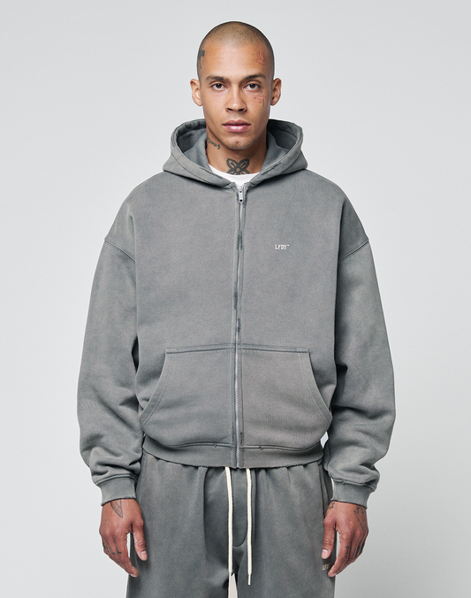 A person in an oversize fit gray Basic Hooded Zip hoodie by LIVE FAST DIE YOUNG, featuring a small white chest logo, front pockets, and matching sweatpants, stands facing forward against a plain light gray background.