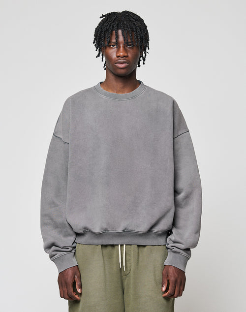A person with short, twisted hair wears a loose grey LIVE FAST DIE YOUNG Basic Blank Sweater and olive green pants, standing against a plain light background and looking directly at the camera with a neutral expression.