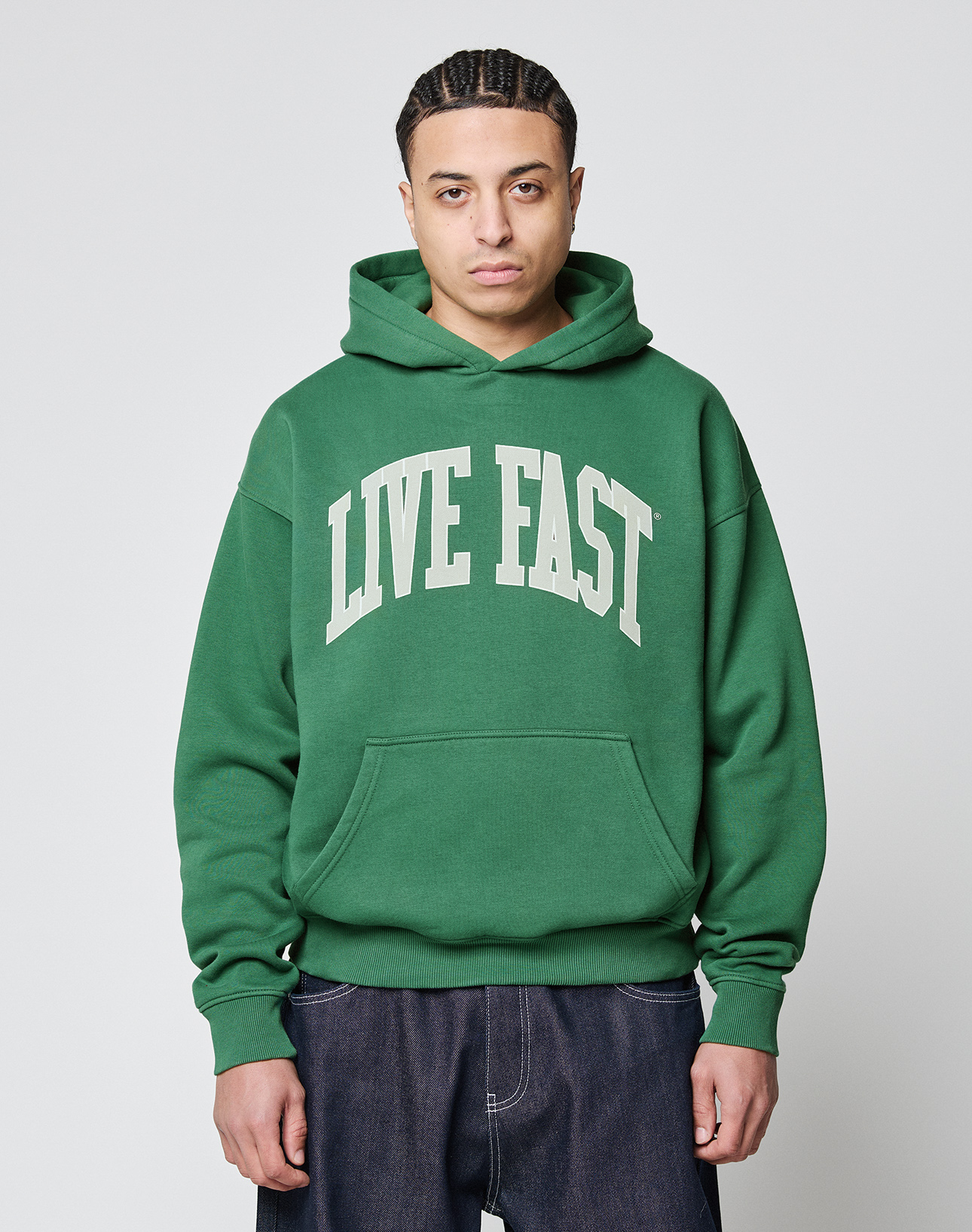 A person wears the LIVE FAST DIE YOUNG Live Fast College Hooded, an oversize fit hoodie in Baumwolle Polyester with a bold white 'LIVE FAST' frontprint, standing against a plain light gray background and looking at the camera.