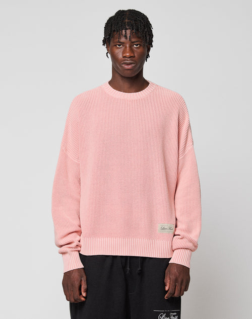 A person with short, textured hair wears the LIVE FAST DIE YOUNG LFDY Knit Sweater in light pink with dropped shoulders and black pants, standing against a plain light gray background.