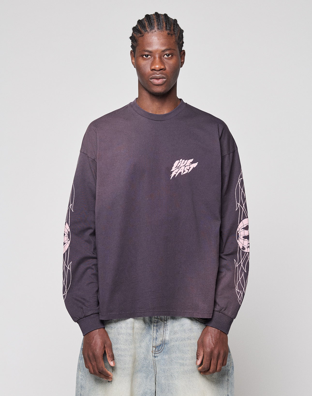 A man with braided hair wears a LIVE FAST DIE YOUNG LFDY Logo Longsleeve—an oversized, 100% cotton dark shirt with white "DIME FAST" on the chest and pink geometric sleeve details, paired with light blue wide-leg jeans.