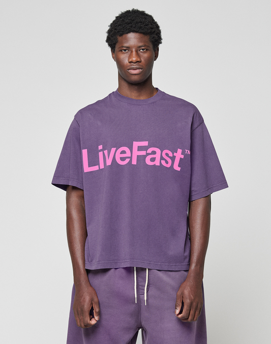 A man faces forward in a LIVE FAST DIE YOUNG Basic OS Livefast Tee, an oversize purple shirt made from 100% Baumwolle with a bold "LiveFast" front print, styled with matching purple sweatpants against a plain light background.