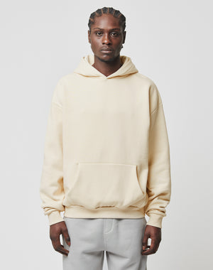 A person with braided hair wears the LIVE FAST DIE YOUNG Basic Blank Hooded cream-colored, oversize hoodie made from soft cotton and polyester, paired with light gray pants, standing against a plain white background.