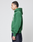 A person with braided hair stands in profile against a plain light background, wearing the LIVE FAST DIE YOUNG Live Fast College Hooded sweatshirt in green with white front print, paired with dark jeans.