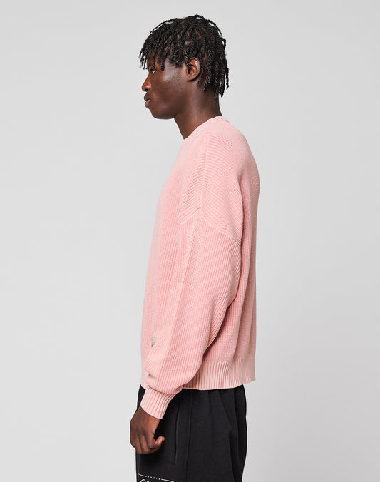 A person with short, twisted hair stands in profile, wearing the LFDY Knit Sweater by LIVE FAST DIE YOUNG and black pants against a plain, light gray background.