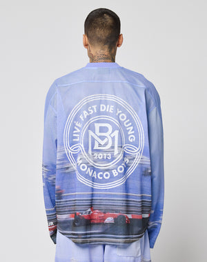 A person with neck tattoos is seen from behind wearing a blue LFDY Yacht Jersey by LIVE FAST DIE YOUNG, featuring an oversize fit, a circular white logo reading LIVE FAST DIE YOUNG MONACO BOYS 2013, and race cars at the bottom.