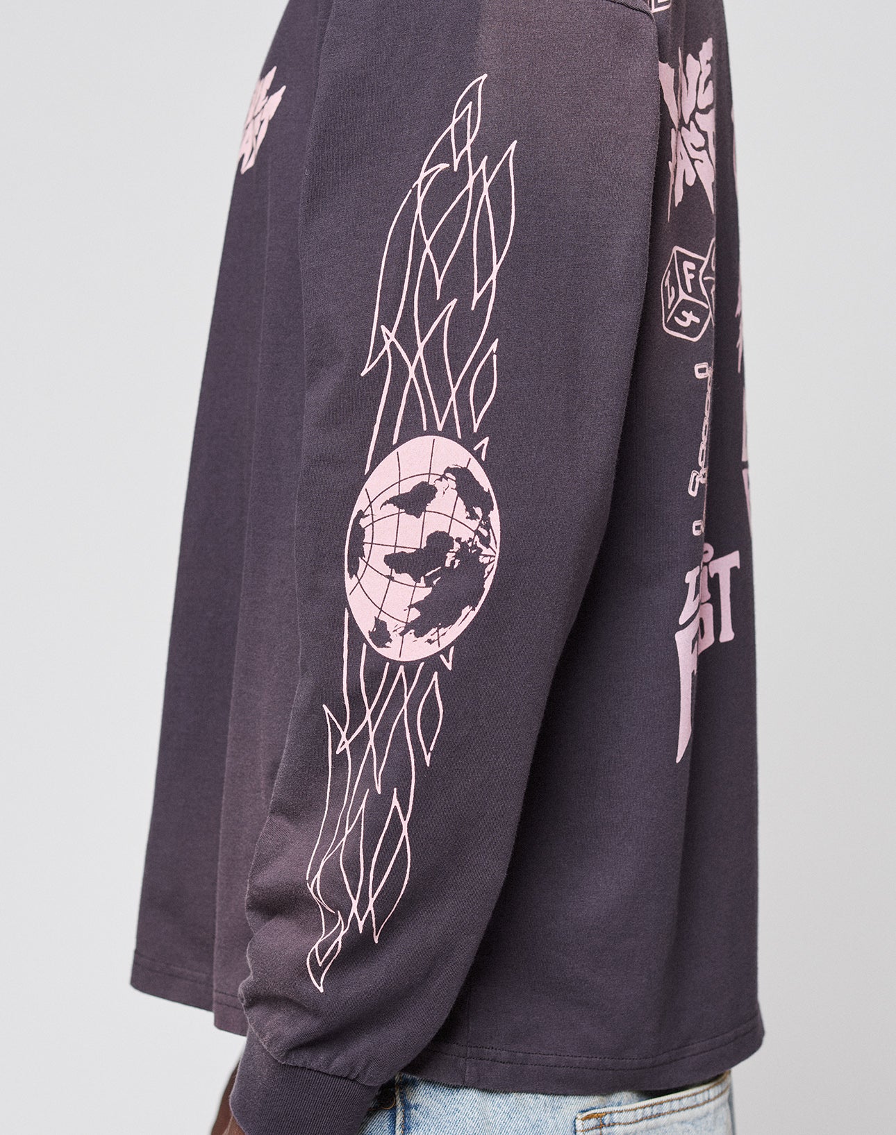 A person wearing the oversize-fit LFDY Logo Longsleeve by LIVE FAST DIE YOUNG, featuring dark fabric with light pink abstract patterns and a circular world map graphic on the sleeve, paired with light blue jeans.