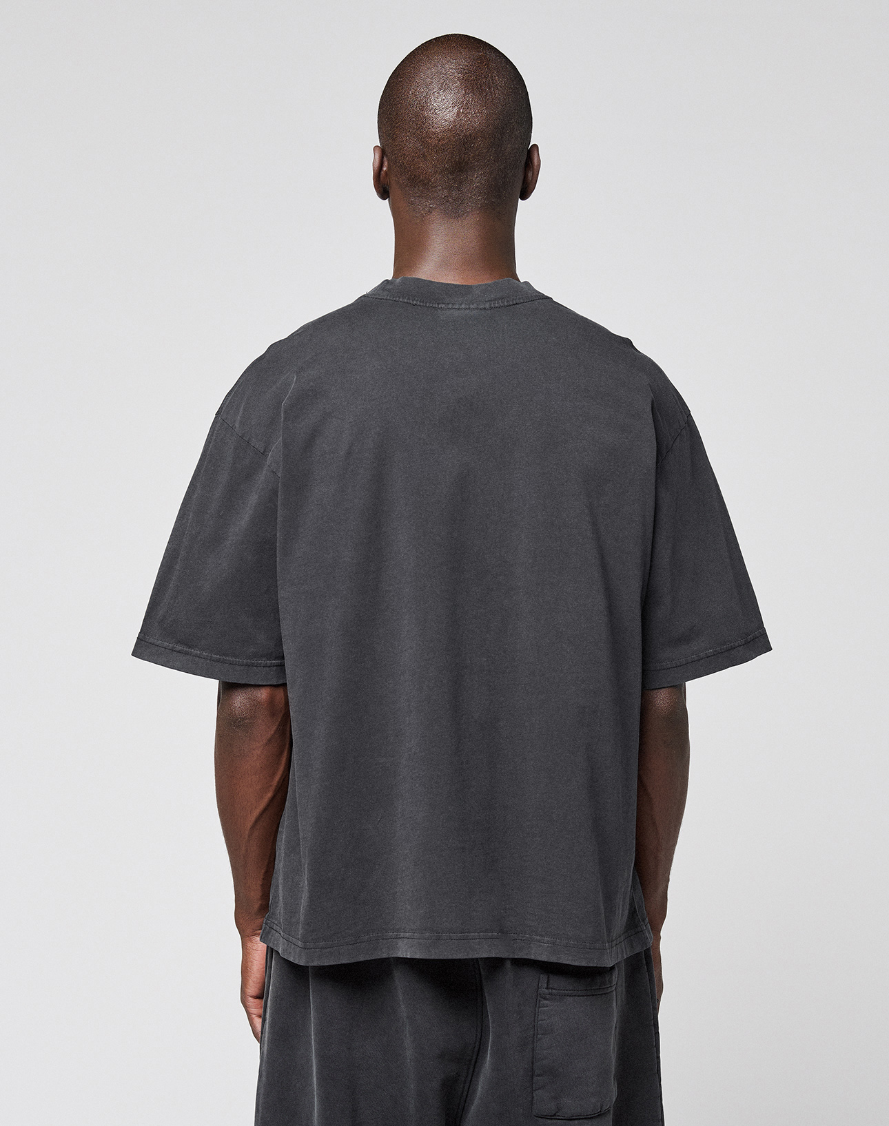 A person with a shaved head faces away from the camera, wearing an oversize fit, dark gray LFDY 40 Tee and matching shorts by LIVE FAST DIE YOUNG, made from 100% Baumwolle, against a plain light gray background.