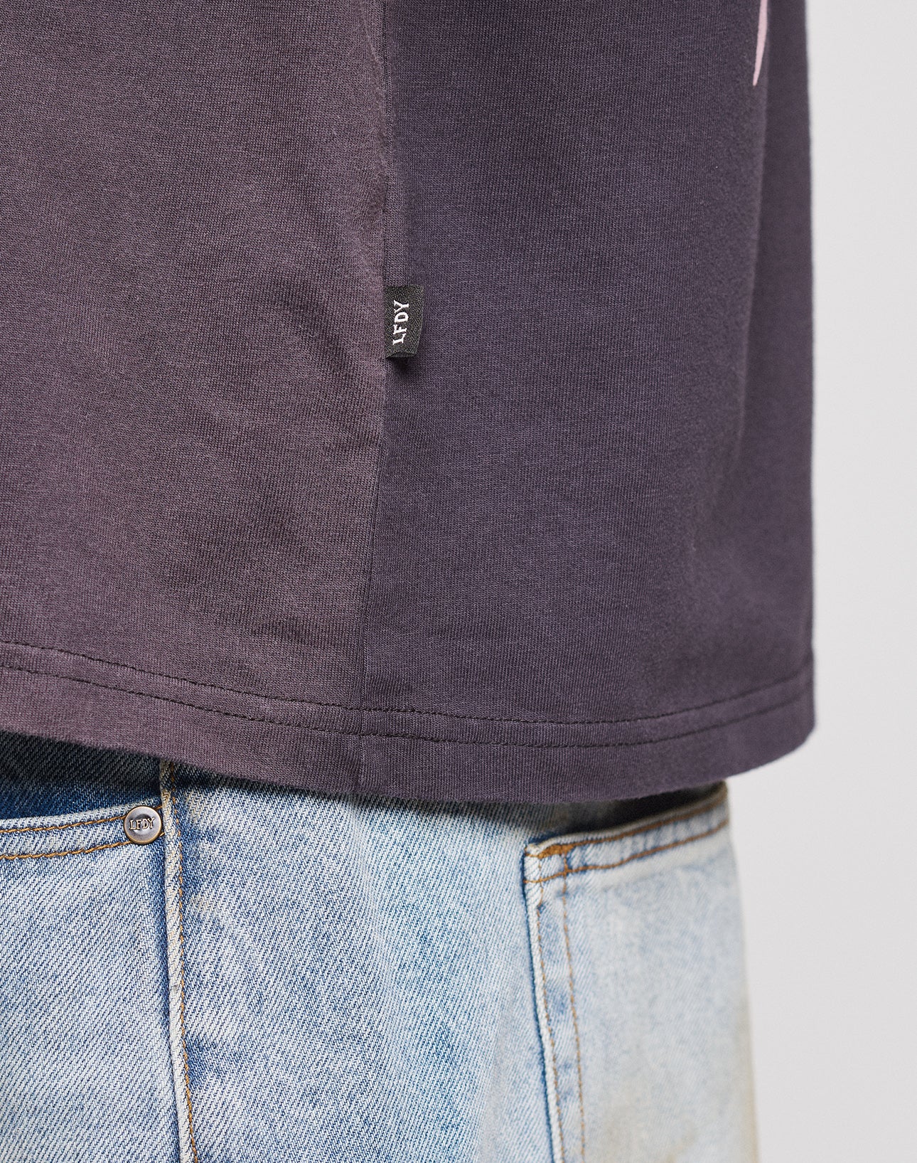 Close-up of the lower side of a dark purple LFDY Logo Longsleeve by LIVE FAST DIE YOUNG, featuring a small black AGM tag, tucked into light blue denim jeans against a plain neutral background.