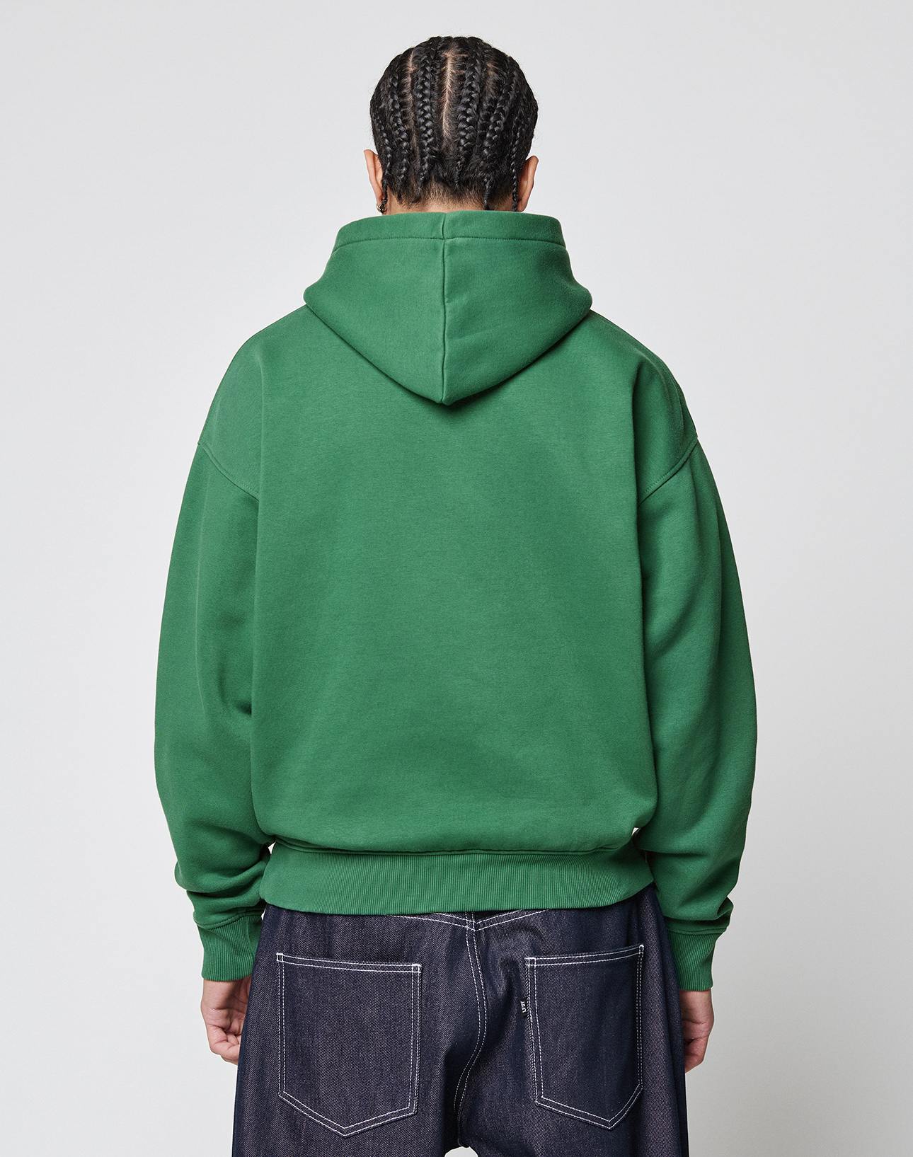 A person with braided hair faces away, wearing the LIVE FAST DIE YOUNG Live Fast College Hooded sweatshirt in green and dark blue jeans against a plain light background.