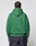 A person with braided hair faces away, wearing the LIVE FAST DIE YOUNG Live Fast College Hooded sweatshirt in green and dark blue jeans against a plain light background.