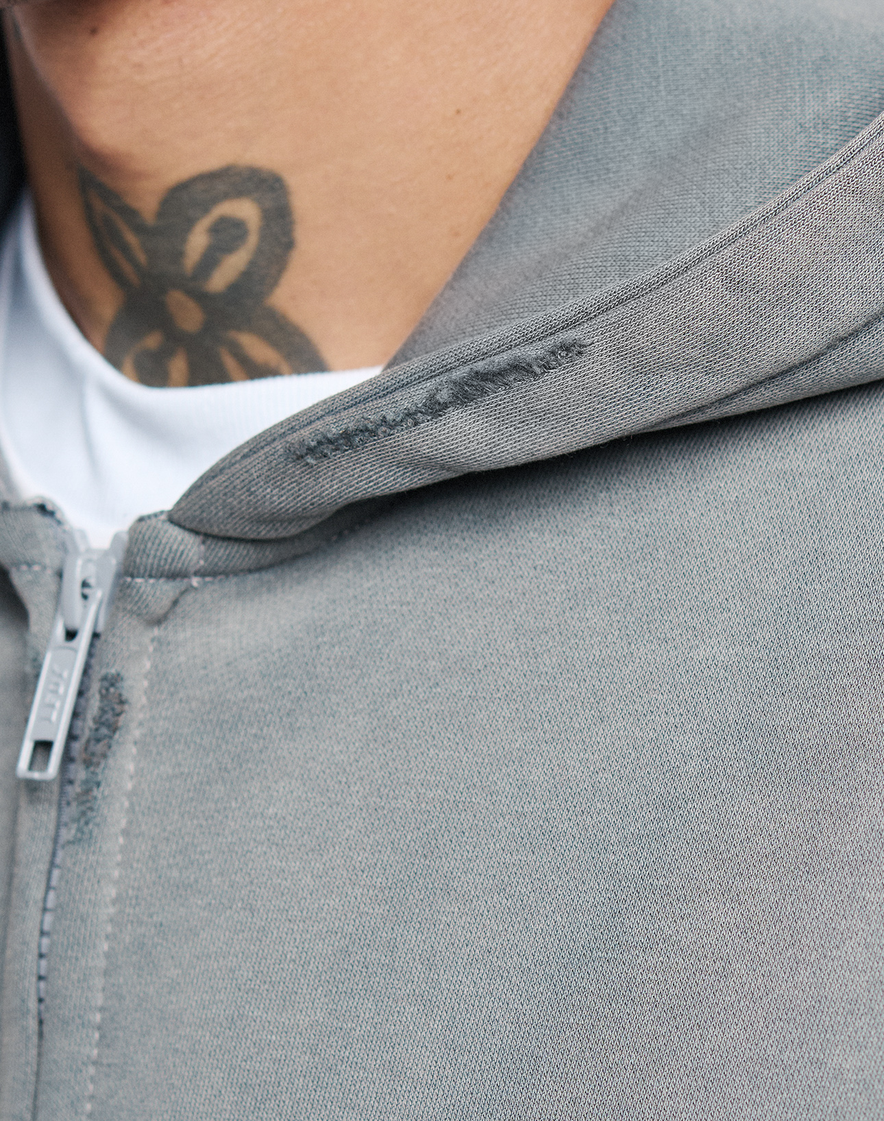 Close-up of a person wearing the LIVE FAST DIE YOUNG Basic Hooded Zip in light gray over a white shirt, revealing part of their neck and a floral or abstract tattoo below the jawline.