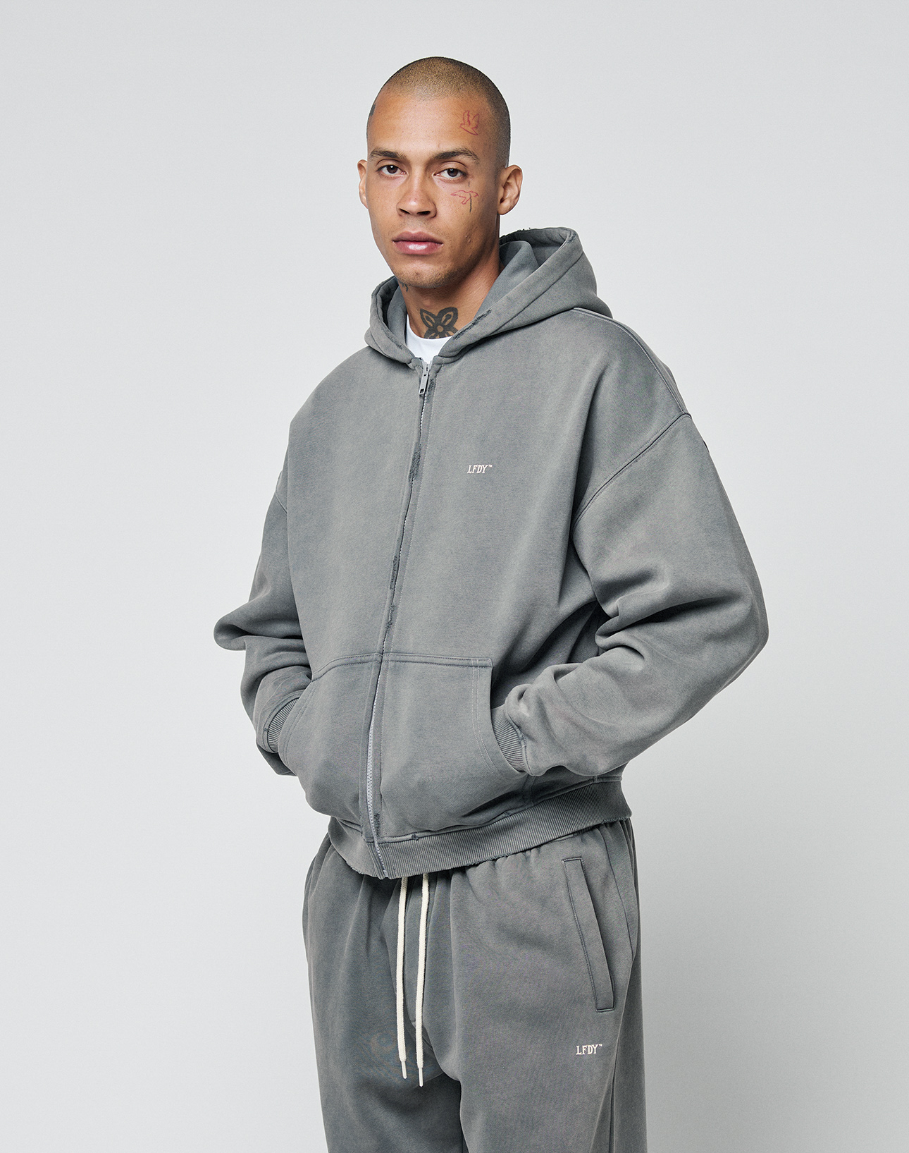 A person wearing an oversize fit Basic Hooded Zip in gray from LIVE FAST DIE YOUNG and matching sweatpants stands against a plain light background, hands in their pockets, with simple neck and face tattoos visible.
