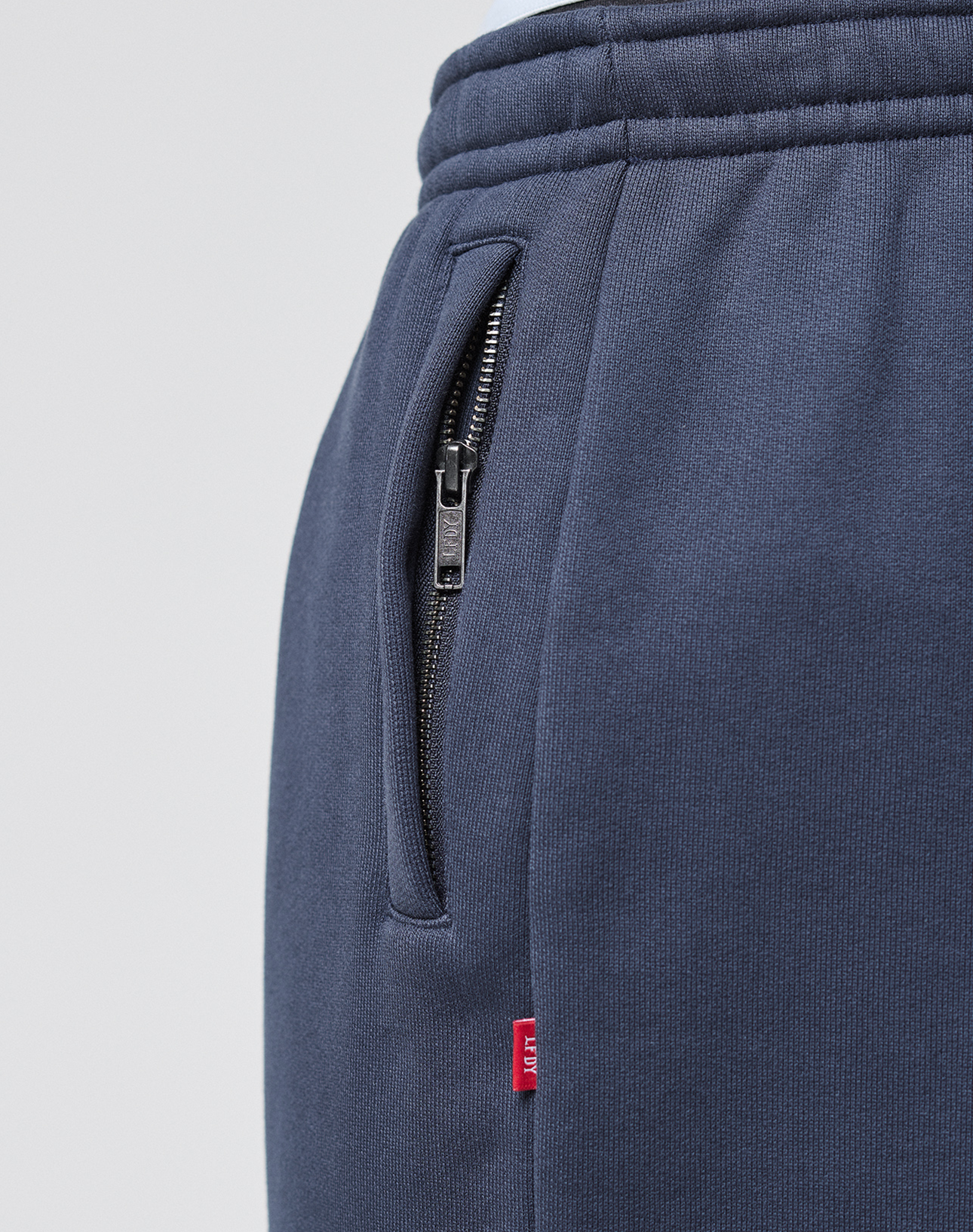 Close-up of the navy blue LFDY Heavyweight Loungepants by LIVE FAST DIE YOUNG, showing a black zipper pocket with a small red fabric tag on the side seam and the clearly visible 650gsm heavyweight fabric texture.