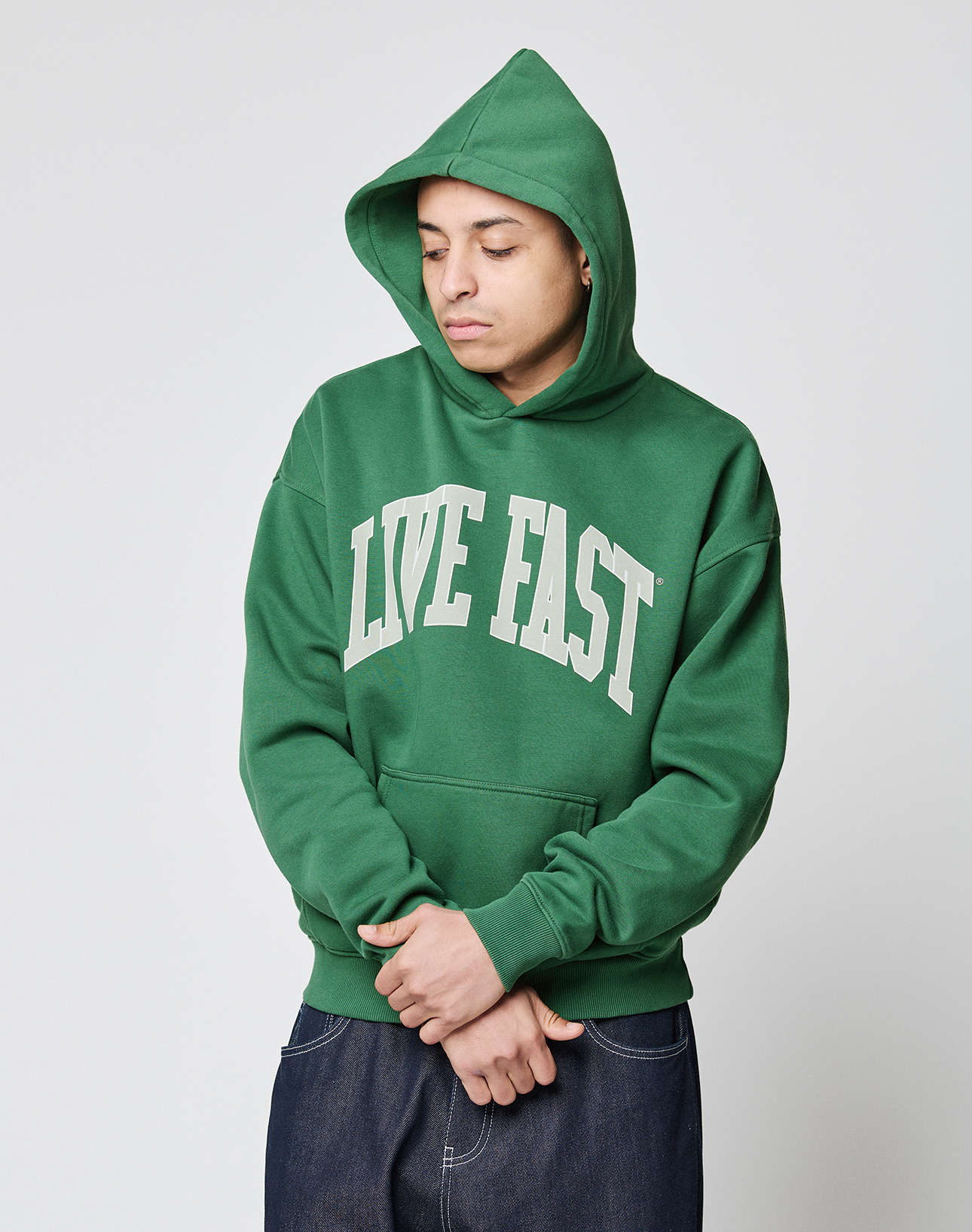 A person in a LIVE FAST DIE YOUNG Live Fast College Hooded green oversize hoodie with front print stands with eyes closed and hands clasped against a plain light background.
