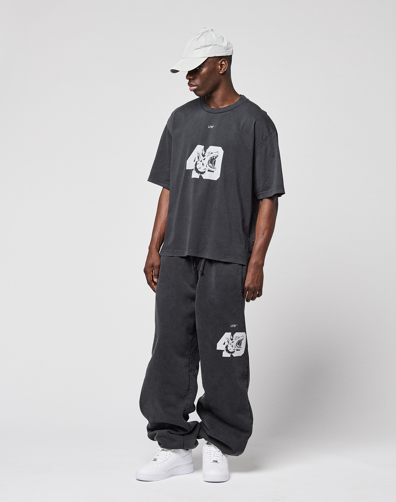 A person in a light gray cap, LFDY 40 Wide Cuff Jogger by LIVE FAST DIE YOUNG, loose dark gray oversized graphic tee, and white sneakers stands against a plain white background.