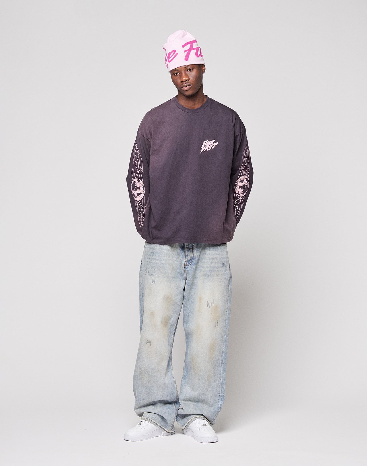 A person wearing the LIVE FAST DIE YOUNG LFDY Logo Longsleeve, pink beanie, light blue baggy jeans, and white sneakers stands against a plain light background.