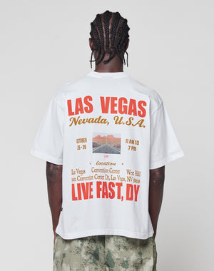 A person faces away, wearing the LIVE FAST DIE YOUNG "Live Fast Vegas Tee"—an oversize fit white cotton shirt with bold red/yellow LAS VEGAS Nevada, U.S.A. prints and event details. Light green patterned pants complete the look.