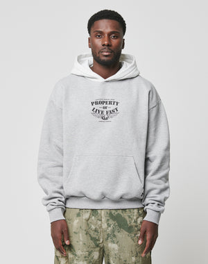 A man in an oversized light gray Live Fast Property Hooded by LIVE FAST DIE YOUNG with chest frontprint and Kapuze, paired with camo pants, stands facing the camera against a plain light background.